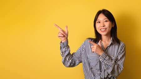 Smiling young Asian woman pointing at blank space, posing and smiling over yellow studio background, banner designの写真素材