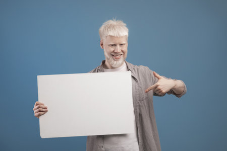 Place for your ad. Excited albino guy holding empty blank board and pointing at it, posing on blue backgroundの写真素材