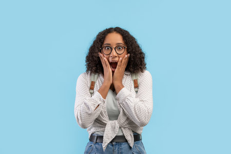Scared young black female student in glasses wearing backpack, screaming in terror on blue studio...