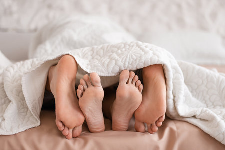 Cropped view of millennial couple lying under blanket on bed, making love at home, closeup of feetの写真素材