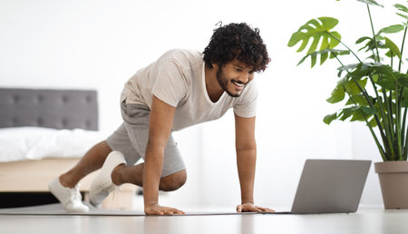 Active hindu guy in sportswear exercising at home, using laptopの写真素材