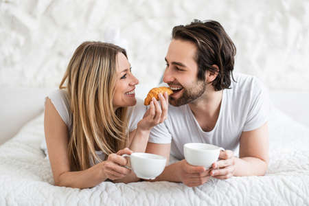 Loving young couple eating breakfast and drinking coffee in bed at home. Lazy romantic morning conceptの写真素材