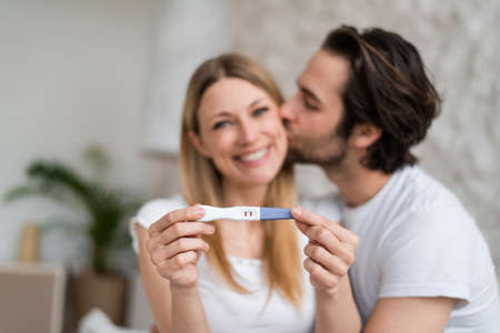 Happy young couple showing positive pregnancy test and kissing at home, selective focusの写真素材