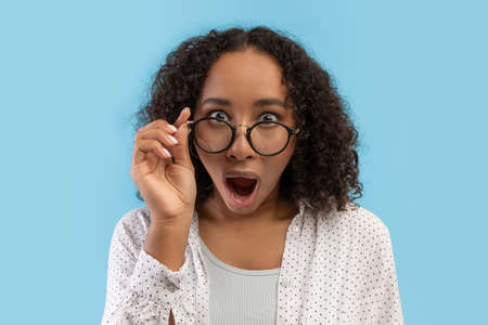 Emotional millennial black female student opening mouth in shock, touching glasses on blue studio backgroundの写真素材