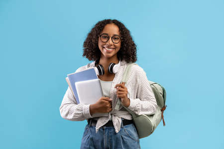 Online education. Smiling black female student with backpack and headphones holding notebooks and...