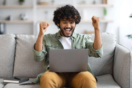 Happy indian man with laptop raising hands up, home interiorの写真素材