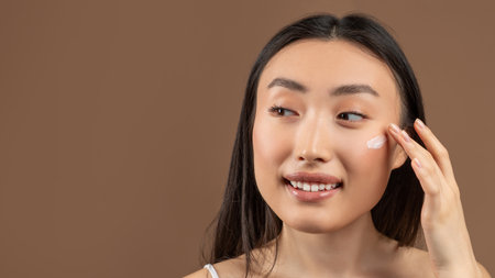 Female skincare concept. Happy asian woman applying moisturizer cream, brown background, copy spaceの写真素材