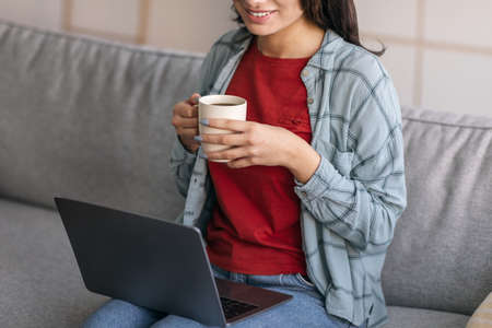 Cropped view of young Arab lady using laptop, working online, watching webinar, drinking coffee at homeの写真素材