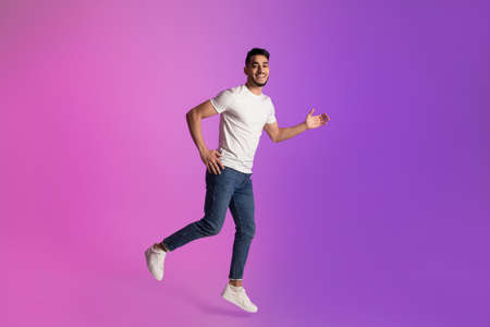 Full length of happy young Arab man in t-shirt and jeans running in neon light, copy space