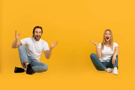 Cheerful surprised young european guy and woman with open mouths raise their hands and sit on floorの写真素材