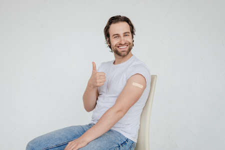 Glad millennial caucasian man in white t-shirt sit on chair, show thumb up and plaster aid on shoulderの写真素材