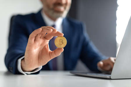 Online Trading. Smiling Businessman In Suit Sitting At Desk And Holding Bitcoinの写真素材