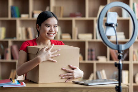 Happy young asian woman hugging cardboard box and look at smartphone webcam in living room