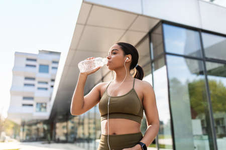 Young black woman in sportswear drinking water, listening to music in earphones during training on...