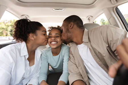 Cheerful Black Parents Kissing Daughter Sitting In New Carの写真素材
