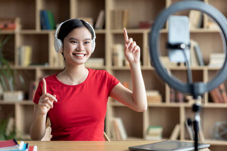 Happy young asian woman in wireless headphones gesturing with hands and look at smartphone webcam