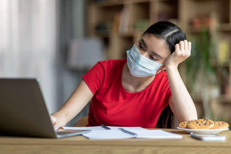 Unhappy young korean lady in protective mask sits at table with laptop in room interior