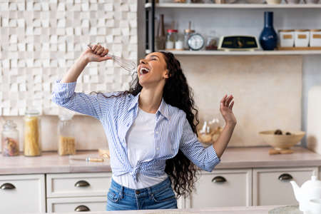 Overjoyed young lady singing at appliance as imaginary microphone, dancing in kitchen interiorの写真素材