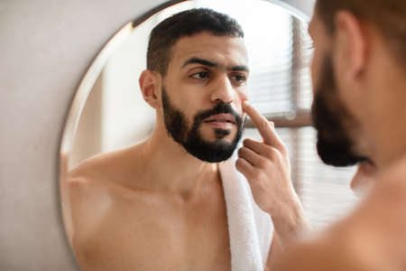 Guy looking in the mirror applying cream on faceの写真素材