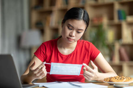 Sad young asian female look at protective mask in hands, sit at table with laptop in living room...