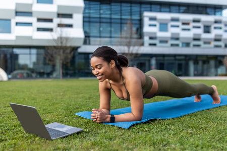 Sporty young black woman standing in plank, training to online sports video on laptop outdoors, copy...