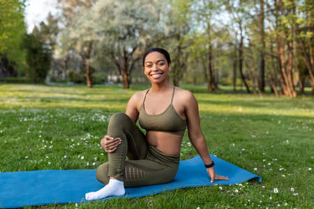 Sporty young black lady stretching legs on yoga mat at summer park, full length