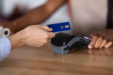 Cropped young black guy bartender in apron at bar counter gives terminal to woman client with credit cardの写真素材