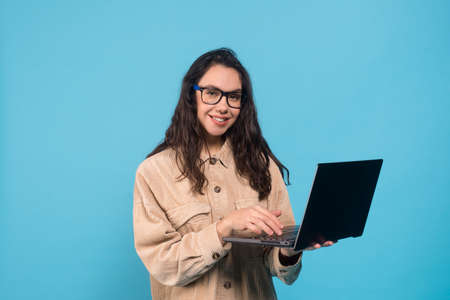 Smiling millennial european woman brunette student in glasses typing on laptop isolated on blue...