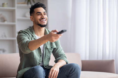 Smiling islamic guy with beard sits on sofa with beard with remote controls watches match on TV,...