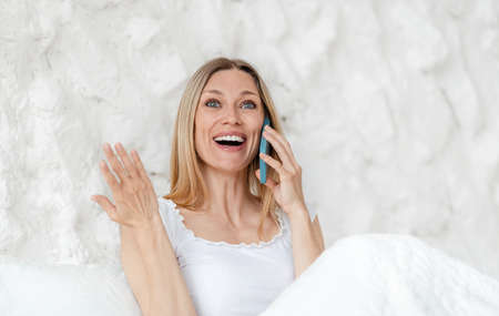 Glad excited pretty young blonde european woman with open mouth speaks by phone in bed in white...