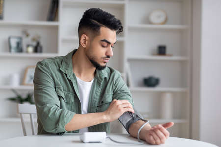 Serious sad young middle eastern man with beard measures blood pressure with tonometer, feels bad