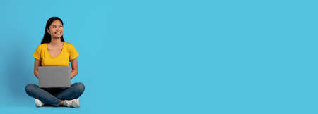 Happy young asian woman in casual sit on floor with laptop, look at empty space isolated on blue...