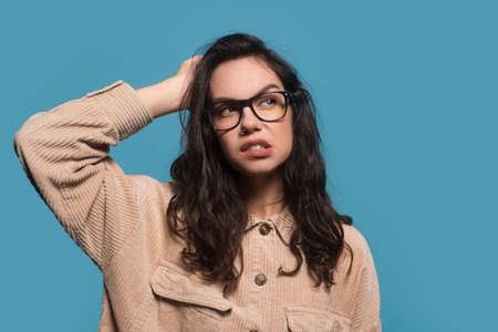 Pensive young european brunette woman in casual and glasses scratches her head and looks at free...