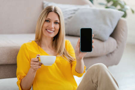 Happy pretty millennial blond european woman in yellow clothes sits on floor, shows phone with empty...