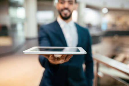 Smiling attractive young arabic male with beard in suit holds tablet with blank screen on his hand...