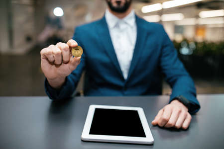 Man in suit with smartwatch hold bitcoin in hand sits at table with tablet in office interiorの写真素材