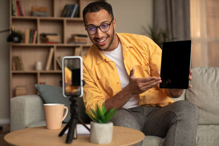 Cheerful black male influencer filming video review of new digital tablet on smartphone at home,...