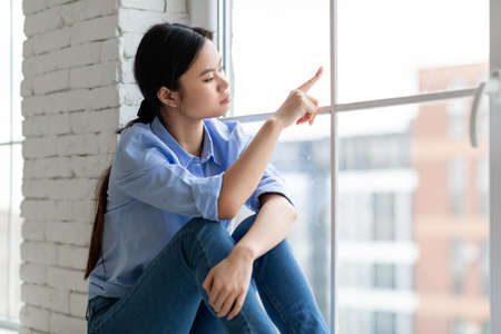 Upset millennial asian woman sitting on window at home