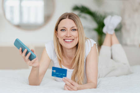 Satisfied pretty young blond caucasian woman with smartphone and credit card lying on bed in white bedroomの写真素材