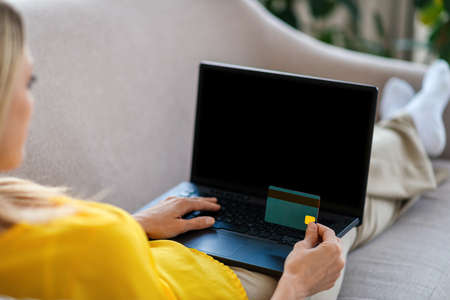 Millennial caucasian lady look at laptop with blank screen, hold credit card, on sofa in living room interiorの写真素材