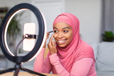 Online makeup tutorial. Happy black woman making video for her internet blog, sitting in front of smartphoneの写真素材