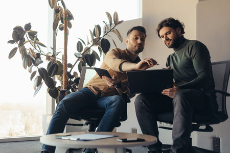 Two male colleagues discussing business project or startup idea, using laptop and digital tablet at modern officeの写真素材