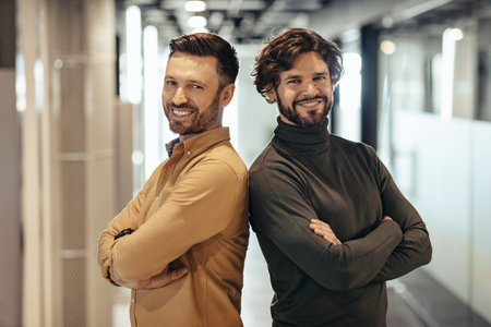 Portrait of two happy businessmen standing with crossed arms, smiling at camera in modern company...