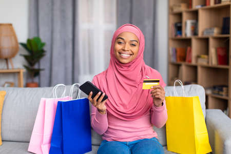 Pretty black muslim woman with shopper bags, credit card and smartphone shopping online from home,...