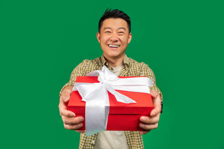 Holiday surprise. Happy middle aged asian man giving gift box to camera and smiling, green studio backgroundの写真素材