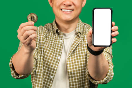 Online trading app. Asian man showing golden bitcoin and smartphone with blank white screen, mockup