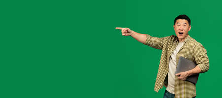 Look there. Surprised asian mature man holding laptop and pointing aside at free space on green background, panoramaの写真素材