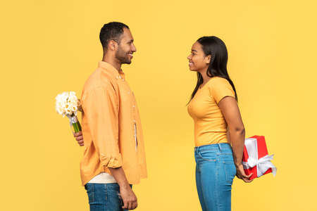 Young black couple hiding gift box and daffodil flowers for each other behind their backs over yellow backgroundの写真素材