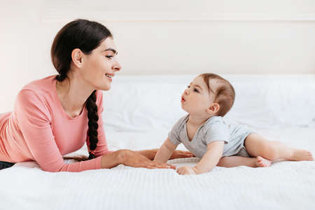 Young mother playing with her cute baby, talking and spending time together, lying on bed in bedroom...