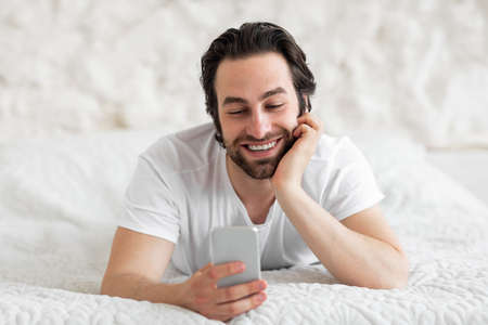 Positive guy chatting with friends on smartphone, lying on bed
